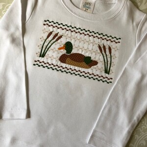 May include: White long-sleeve shirt with an embroidered design of a duck in a marsh. The shirt has a label that reads "GRB Blanks 2T".