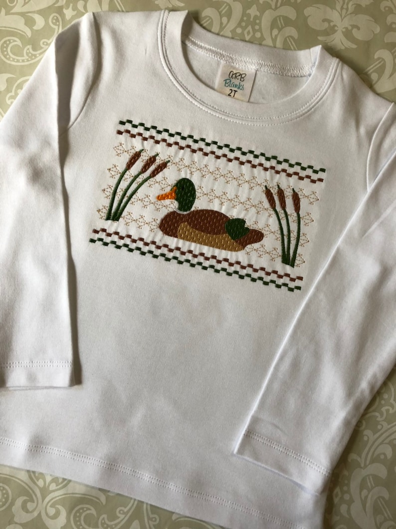 May include: White long-sleeve shirt with an embroidered design of a duck in a marsh. The shirt has a label that reads "GRB Blanks 2T".