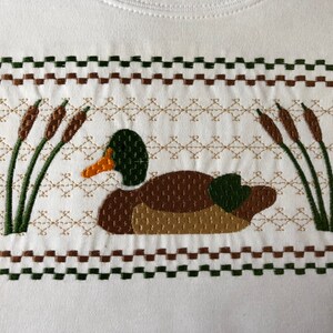 May include: Embroidered design of a brown duck with green head and wings, swimming in a pond with green reeds. The design is surrounded by a brown and green checkered border.
