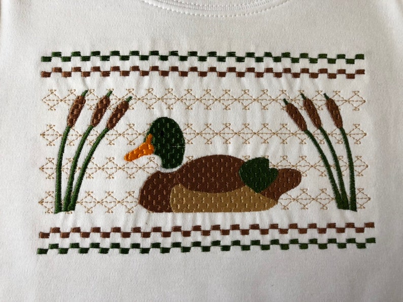 May include: Embroidered design of a brown duck with green head and wings, swimming in a pond with green reeds. The design is surrounded by a brown and green checkered border.