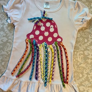 May include: White t-shirt with a pink and white jellyfish applique. The jellyfish has a smiling face and rainbow colored ribbon tentacles. The shirt has a ruffled hem and says "CCB Blanks" on the tag.