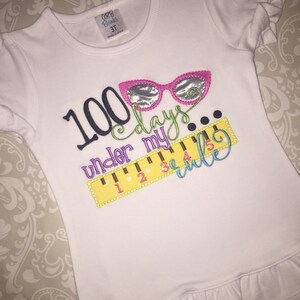 May include: White t-shirt with a colorful embroidered design that says "100 days under my rule" with a ruler and glasses.