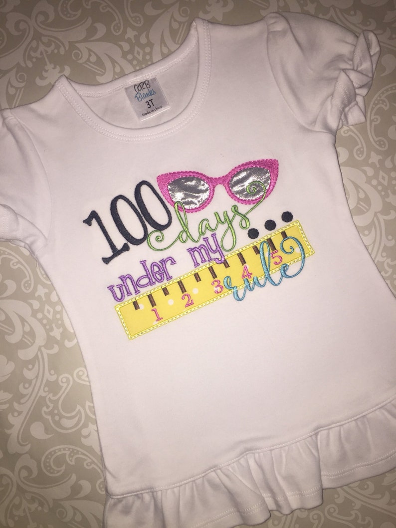 May include: White t-shirt with a colorful embroidered design that says "100 days under my rule" with a ruler and glasses.