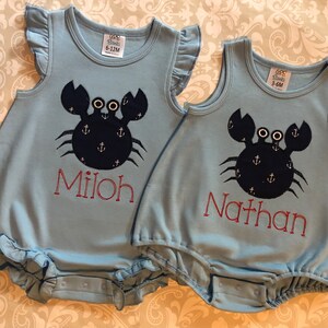 May include: Two light blue baby onesies with a black crab design and anchor accents. The onesies have the names "Milo" and "Nathan" embroidered on them in red.