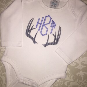May include: White long-sleeve baby onesie with a monogram of the letters "HBT" embroidered in purple on a gray antler design. The onesie has a label that reads "CRB Blanks 0-3M".