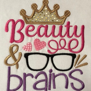 May include: Embroidered design on a white t-shirt featuring a gold crown, pink heart, and the text "Beauty & Brains" in pink, gold, and purple.