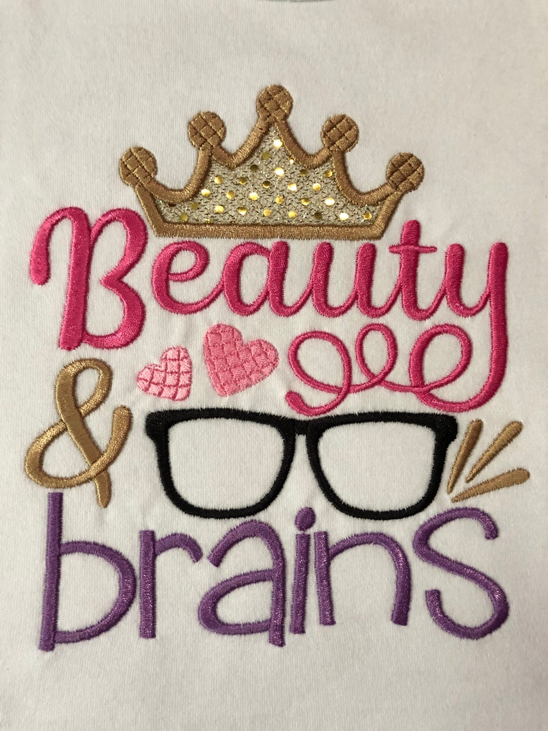 May include: Embroidered design on a white t-shirt featuring a gold crown, pink heart, and the text "Beauty & Brains" in pink, gold, and purple.