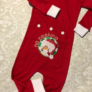 May include: Red Christmas onesie with white trim and a snowman embroidered on the back pocket. The snowman is wearing a red hat and has the words "no peeking" written around it.