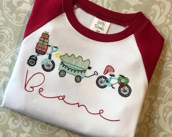 May include: White and red raglan-style shirt with an embroidered design of a Christmas tree, presents, and a bicycle. The name "Beane" is embroidered below the design.