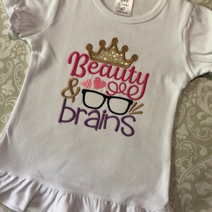 May include: White t-shirt with a pink, purple, and gold embroidered design that says "Beauty & Brains" with a crown and glasses. The shirt has a ruffled hem and is size 4T.