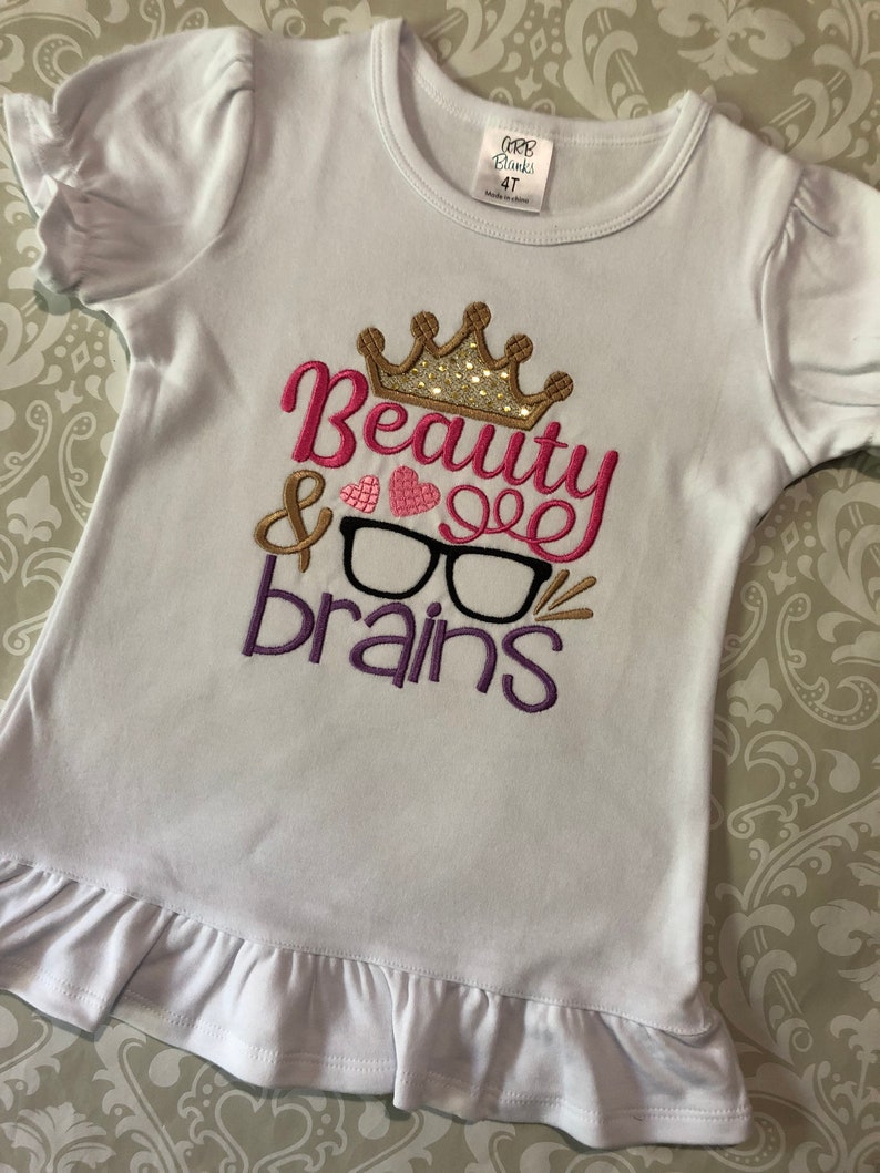 May include: White t-shirt with a pink, purple, and gold embroidered design that says "Beauty & Brains" with a crown and glasses. The shirt has a ruffled hem and is size 4T.