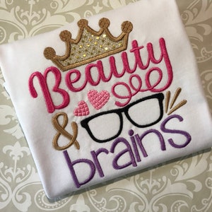 May include: White t-shirt with pink, gold, and purple embroidery. The embroidery says "Beauty & Brains" with a crown, heart, and glasses.