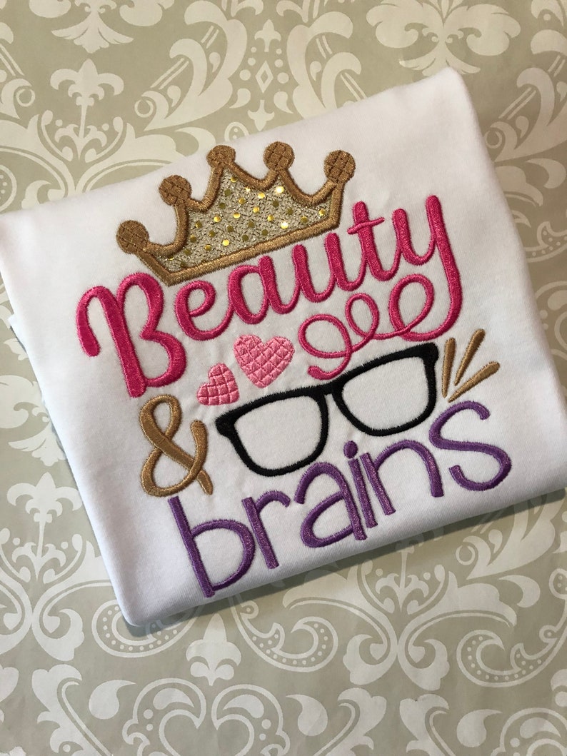 May include: White t-shirt with pink, gold, and purple embroidery. The embroidery says "Beauty & Brains" with a crown, heart, and glasses.