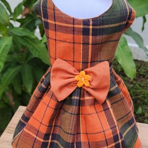May include: A plaid dog dress in orange, green, and navy blue with a matching bow and headband. The dress features a large bow with a small orange flower accent. The fabric has gold metallic threads.