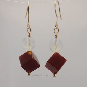 May include: Dangle earrings with gold-coloured hooks. Each earring has a clear, round bead, a faceted, reddish-brown cube-shaped bead, and a small gold-coloured bead at the bottom. The earrings are suspended against a neutral background.