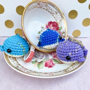 May include: Three crocheted whale plushies in blue, purple, and white with black eyes. The plushies are sitting on a white teacup and saucer with a floral pattern.