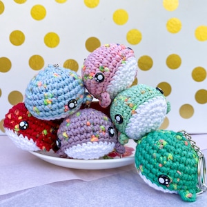 May include: A set of six crocheted whale keychains in various colors, including blue, pink, green, purple, and red. Each whale has a white belly and black eyes. The keychains are arranged on a white plate with a floral design.