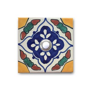 May include: A white ceramic doorbell button with a blue, green, red, and yellow geometric pattern. The button is round and has a silver ring around it.