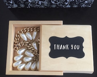 May include: A wooden box with a hinged lid. The box is open and contains a gold chain necklace with white pearl drops. The lid has a black chalkboard label with the text "THANK YOU" written in white.
