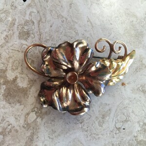 May include: A silver and gold tone flower brooch with a textured finish. The brooch has a leaf design and a pin on the back.