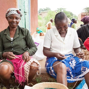 May include: Four women are sitting outside, working on crafts. They are using string to create woven items. The women are wearing colorful clothing and head coverings.