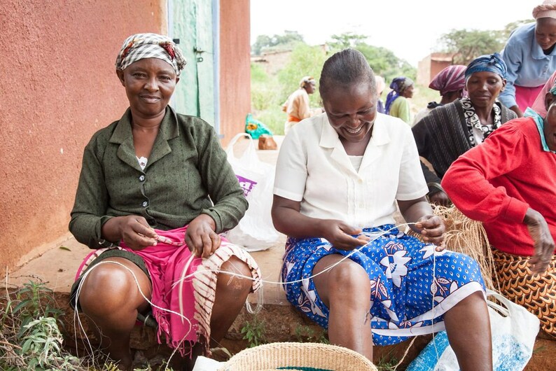 May include: Four women are sitting outside, working on crafts. They are using string to create woven items. The women are wearing colorful clothing and head coverings.