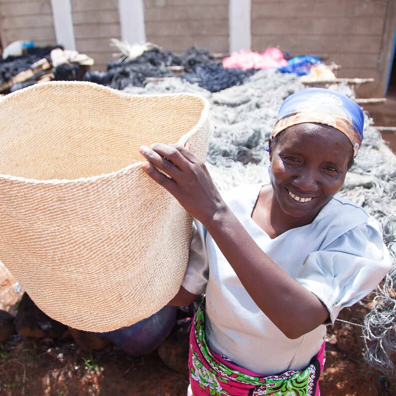 May include: A woman holds a large, woven basket made of natural fibers. The basket is light brown and has a smooth, textured surface. The woman is wearing a blue headscarf and a light blue shirt. She is smiling and looking at the camera.