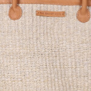 May include: A close-up of a beige woven tote bag with brown leather handles and a brown leather tag that reads "THE BASKET ROOM".
