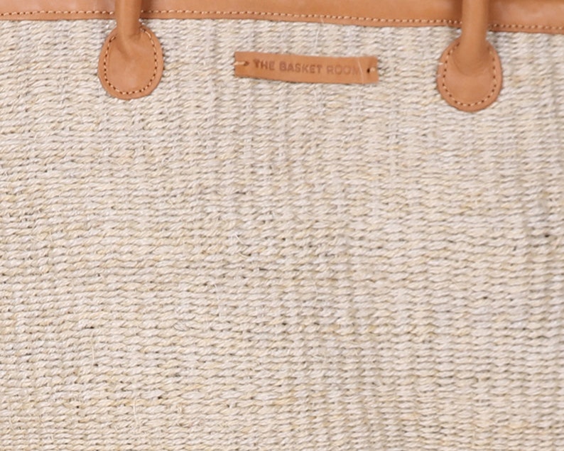 May include: A close-up of a beige woven tote bag with brown leather handles and a brown leather tag that reads "THE BASKET ROOM".