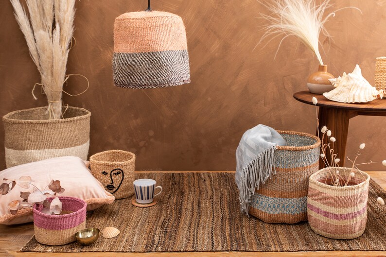 May include: A collection of woven baskets in various sizes and colors, including pink, brown, and blue, with a pink pillow and a seashell on a woven rug.