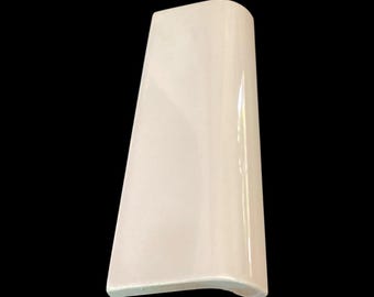 May include: A single, off-white ceramic tile with a curved edge and a glossy finish. The tile is rectangular with a slightly trapezoidal shape, and is set against a black background.