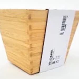 May include: A light brown, square bamboo container with a flared design. A white label with black text and a barcode is wrapped around the side. The container is empty.