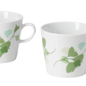 May include: Two white ceramic mugs with a curved handle. Each mug is decorated with a green and light green leaf design. The mugs are set against a white background.