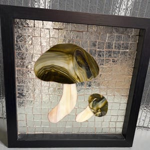 May include: A framed mosaic art piece featuring a mushroom design. The mushroom has a brown and cream-colored cap and a cream-colored stem. The background is a mosaic of small, square, silver tiles, and the frame is black.