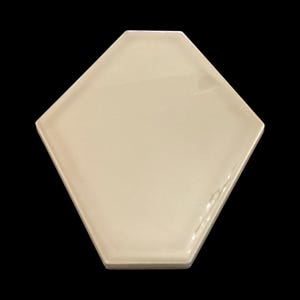 Puede incluir: Una sola baldosa en forma de diamante de color crema con un acabado brillante. La baldosa tiene un diseño geométrico y está colocada sobre un fondo negro. La forma de la baldosa es un hexágono modificado.