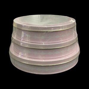 Puede incluir: Un juego de tres latas redondas apilables en color rosa claro. Cada lata presenta un patrón ondulado blanco repetido y un borde dorado. Las latas están envueltas en plástico transparente, lo que sugiere que son nuevas y sin usar.