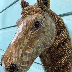 Op de afbeelding: Een mozaïekpaardenkopensculptuur met bruine en gouden glazen tegels. De manen van het paard zijn gemaakt van kleine gouden kralen.
