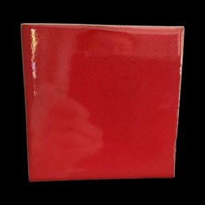 Op de afbeelding: Een vierkant, glanzend, rood object met een glad oppervlak. De randen zijn licht afgerond en de kleur is een levendig, effen rood. Het object staat tegen een zwarte achtergrond.