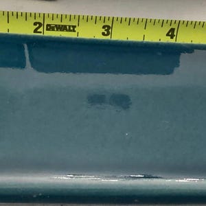 Könnte beinhalten: Eine rechteckige, teal-farbene Fliese mit glänzendem Finish. Die Fliese ist etwa 15 cm lang, gemessen mit einem gelben Maßband. Die Kanten der Fliese sind leicht abgerundet.