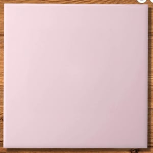 Puede incluir: Una baldosa cuadrada de color rosa pálido con una superficie lisa. La baldosa está colocada sobre un fondo de madera, creando un contraste de texturas y colores. La imagen es una vista superior.