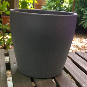 Puede incluir: Una maceta gris oscuro, de forma ovalada. La maceta tiene una superficie texturizada y una parte superior ligeramente ensanchada. Está sobre una superficie de madera, con vegetación visible en el fondo. Ideal para plantas.
