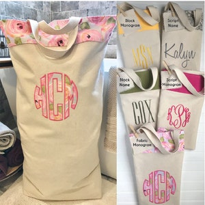 May include: Cream-colored canvas laundry bag with a floral rose pattern trim and handles. The bag features a large embroidered monogram in pink and floral fabric. Additional bags with monograms and names are also shown.