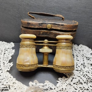 May include: Vintage opera glasses with a mother-of-pearl finish and brass detailing. The binoculars are in a brown leather case with a handle. The glasses have a cream-coloured top and bottom. The case has a gold-coloured button.