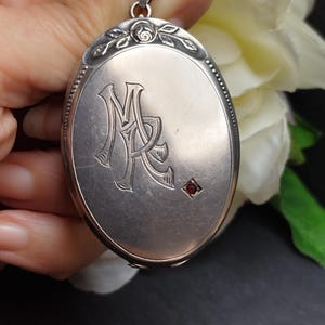 Puede incluir: Medallón ovalado de plata con un monograma grabado "M&R" y un pequeño acento de piedra roja. El medallón tiene un diseño floral alrededor de la parte superior.