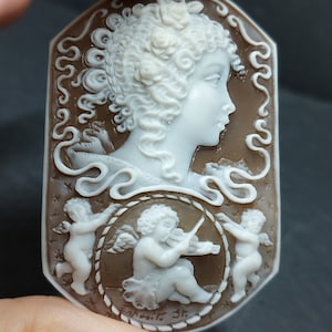 Cameo rettangolare con bordi arrotondati in conchiglia fondo scuro marrone inciso a rilievo colore bianco profilo donna con capelli ricci incisi nei minimi dettagli, circondata da 3 angeli Puttini uno suona il violino ,molto dettagliata l'incisione