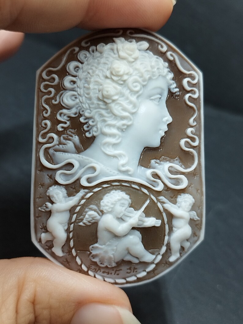 Cameo rettangolare con bordi arrotondati in conchiglia fondo scuro marrone inciso a rilievo colore bianco profilo donna con capelli ricci incisi nei minimi dettagli, circondata da 3 angeli Puttini uno suona il violino ,molto dettagliata l'incisione