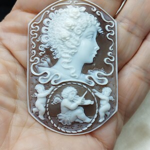 May include: A cameo pendant featuring a carved white woman's profile with curly hair and a flower in her hair. The cameo is set in a brown shell with a rope border and two cherubs, one playing a violin. The cameo is signed "Esposito 3r."