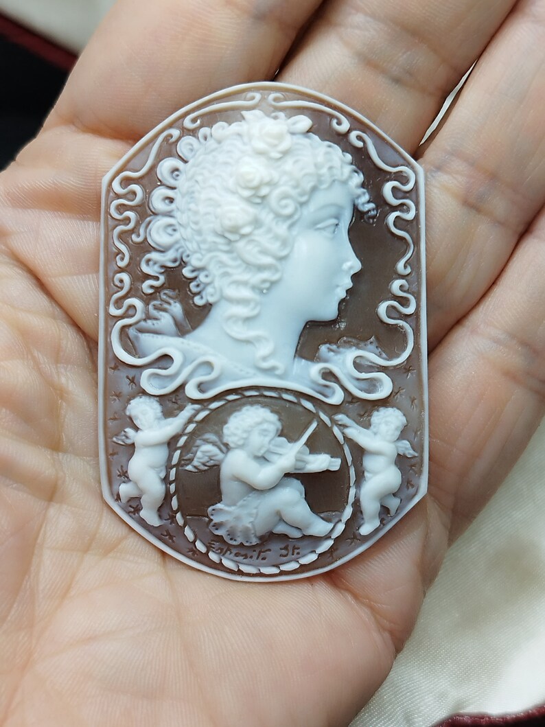 May include: A cameo pendant featuring a carved white woman's profile with curly hair and a flower in her hair. The cameo is set in a brown shell with a rope border and two cherubs, one playing a violin. The cameo is signed "Esposito 3r."