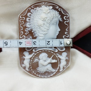 May include: A cameo carved from shell with a brown background and white figures. The cameo features a profile of a woman with flowers in her hair and two cherubs, one playing a violin. The cameo is approximately 7.6 cm long.