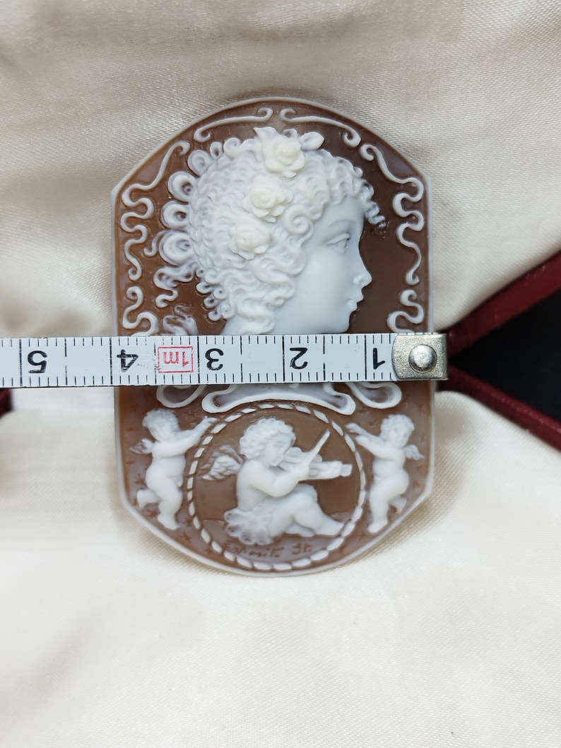 May include: A cameo carved from shell with a brown background and white figures. The cameo features a profile of a woman with flowers in her hair and two cherubs, one playing a violin. The cameo is approximately 7.6 cm long.